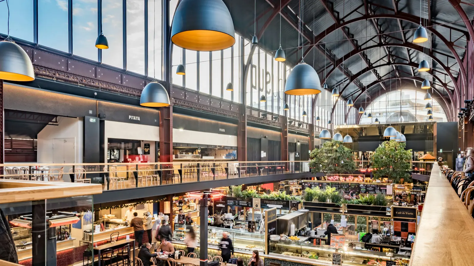 Innenansicht einer modernen Markthalle mit hohen Fenstern, hängenden Lampen und mehreren Essensständen, an denen Menschen sitzen und essen.