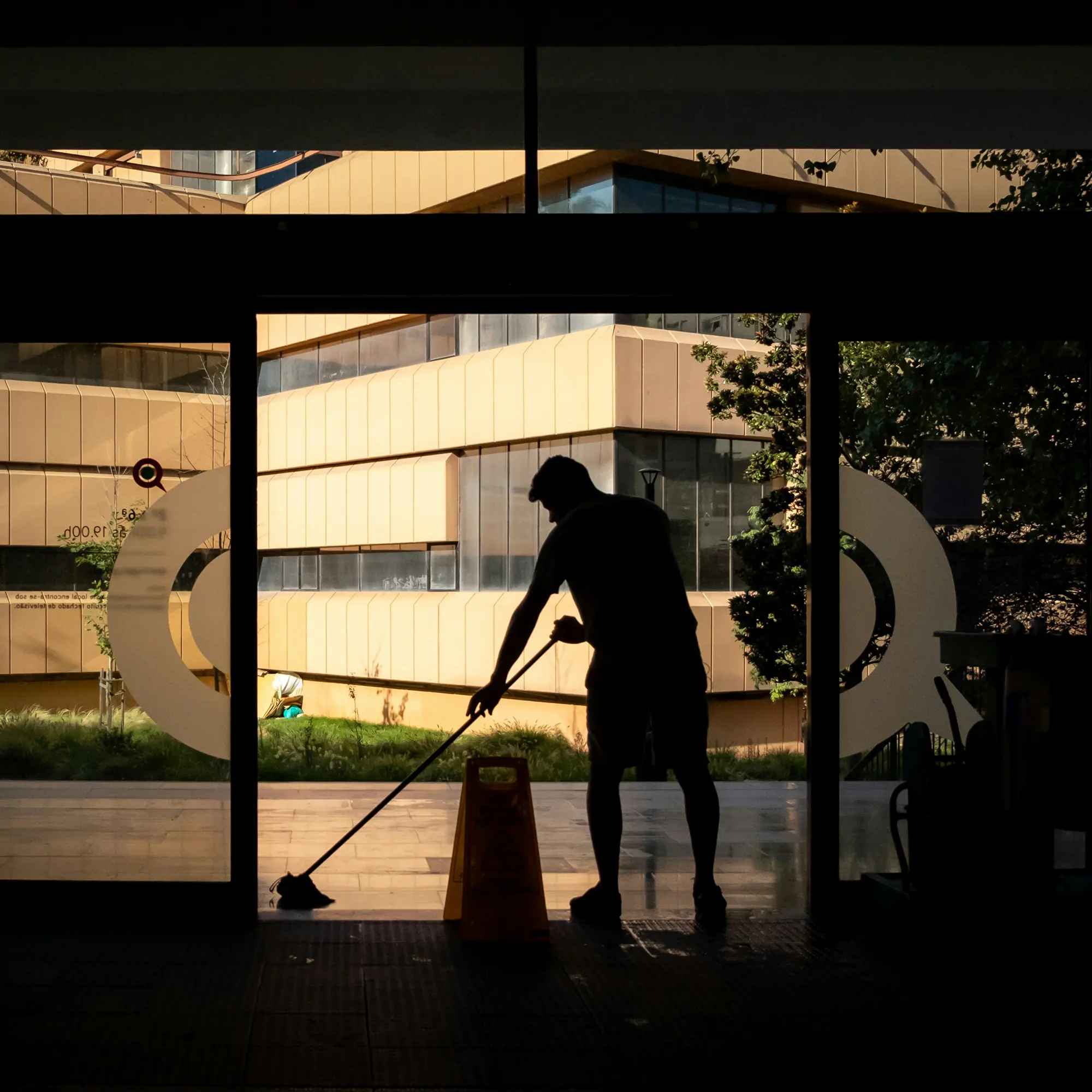 Silhouette einer Person, die den Boden in einem Gebäude mit einem Mopp reinigt, mit hellen Gebäudefassaden im Hintergrund.